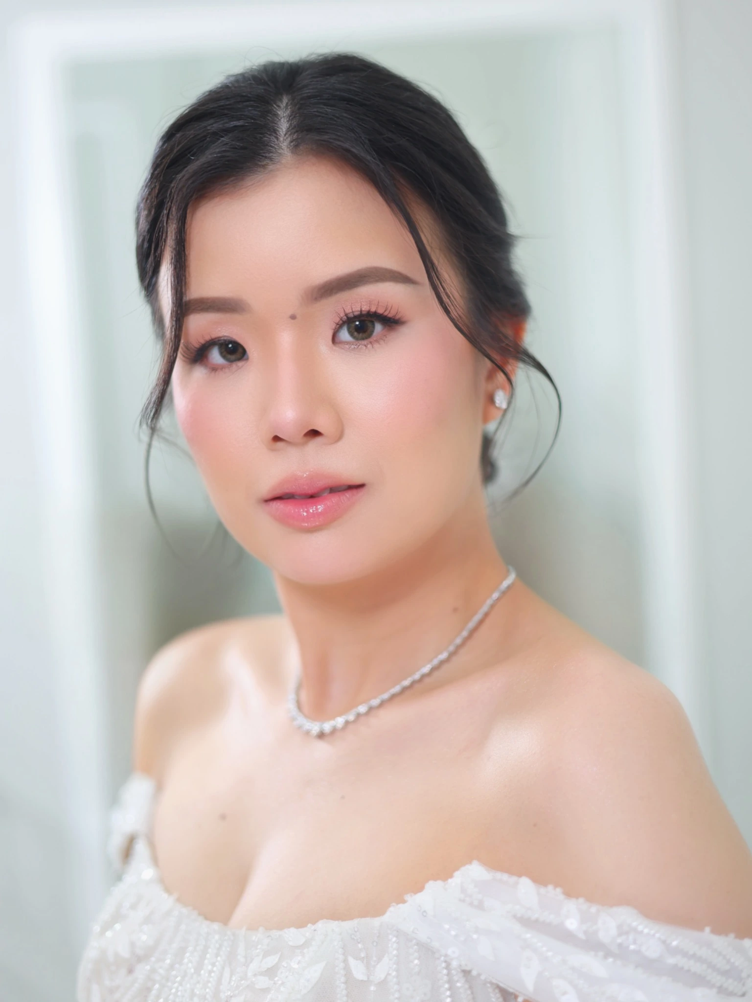 Bridal portrait with a softly styled updo, defined lashes, pink blush and glossy pink lips, wearing a white off-the-shoulder gown and a sparkling necklace