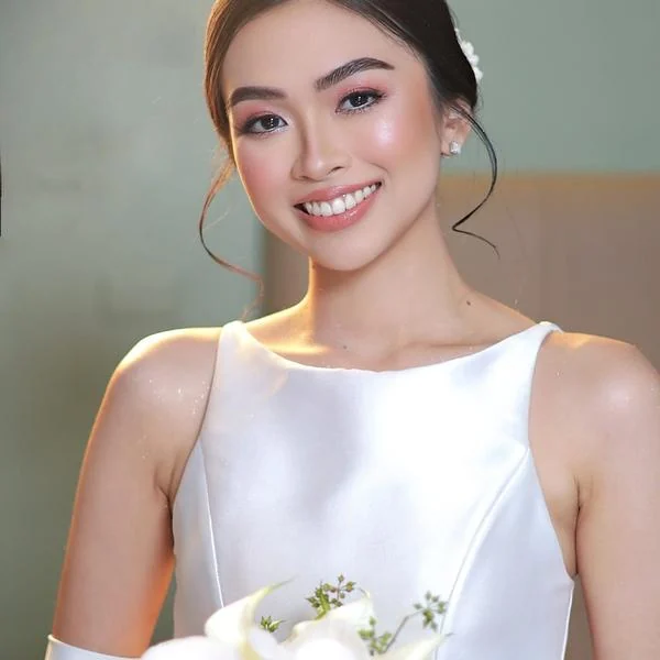Smiling woman holding white flowers, wearing a white dress and rosy pink makeup