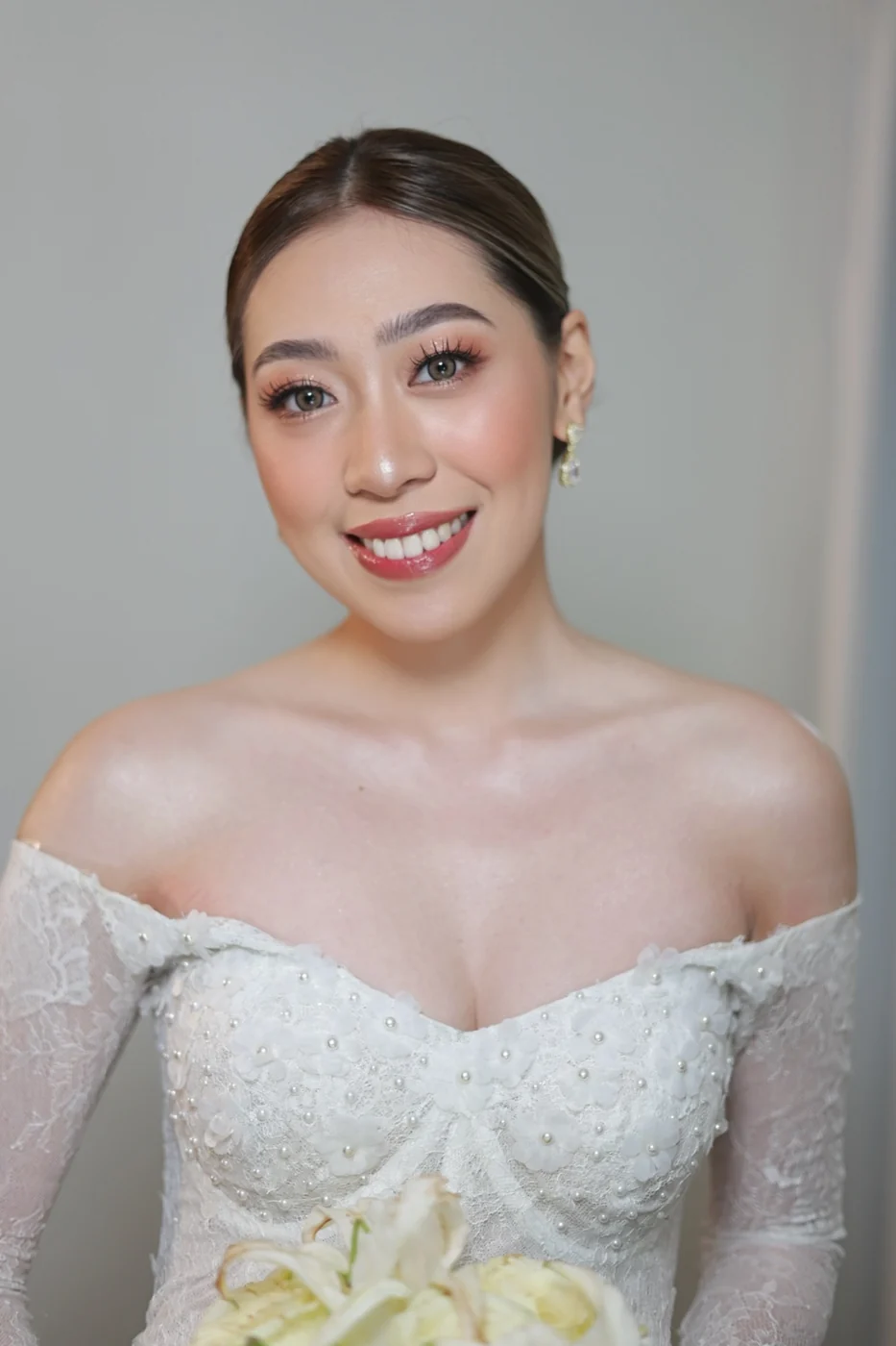 Smiling bride with a sleek center-parted hairstyle, rosy makeup and an off-the-shoulder beaded white gown, holding white flowers