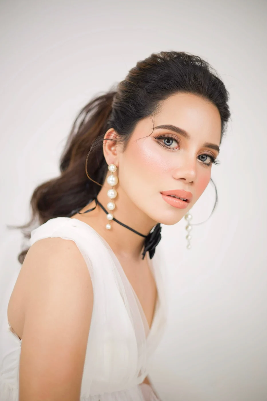 Beauty portrait with a voluminous ponytail, peach-toned makeup, long pearl earrings and a white top